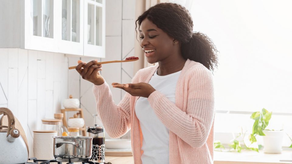 Cheerful Black Housewife Tasting Food While Cooking Healthy Lunch In Modern Kitchen